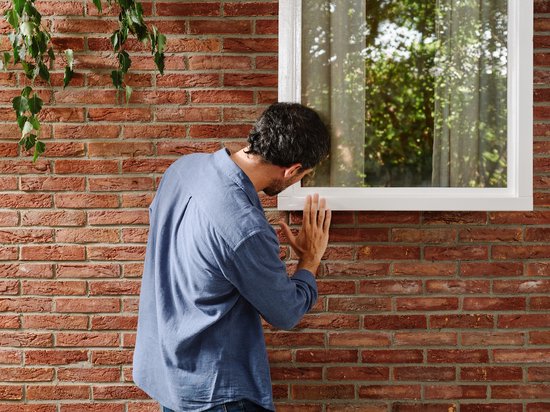 Man in blauw shirt inspecteert een beschadigd wit kozijn naast een bakstenen muur.