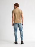 Man met krullend rood haar, gedraaid naar links. Draagt een lichtbruin t-shirt en spijkerbroek.