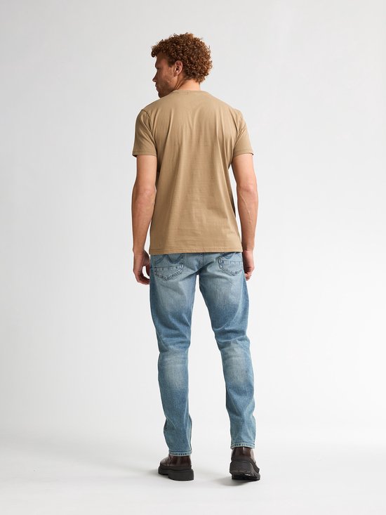 Man met krullend rood haar, gedraaid naar links. Draagt een lichtbruin t-shirt en spijkerbroek.