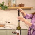 Vrouw met paarse jas gebruikt een zwarte Vacu Vin wijnstopper op een fles rode wijn.