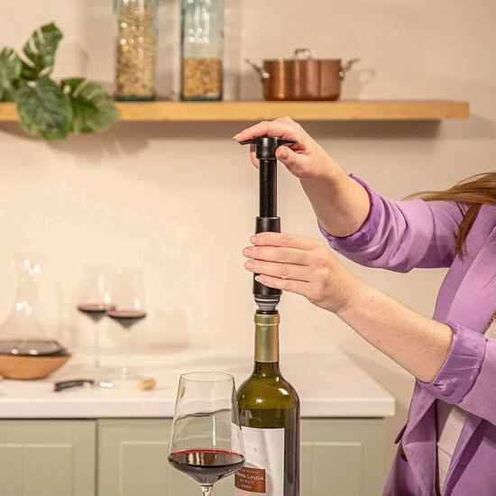 Vrouw met paarse jas gebruikt een zwarte Vacu Vin wijnstopper op een fles rode wijn.