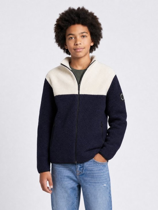 Model in een donkerblauw en off-white teddyjack met rits, handen in de zakken, dragend een spijkerbroek.