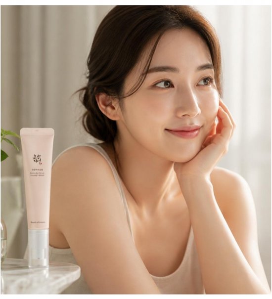 Model met een zachte glimlach houdt haar gezicht vast met haar hand, met een roze tube van Beauty of Joseon Ginseng + Retinol Revive Eye Serum ernaast.