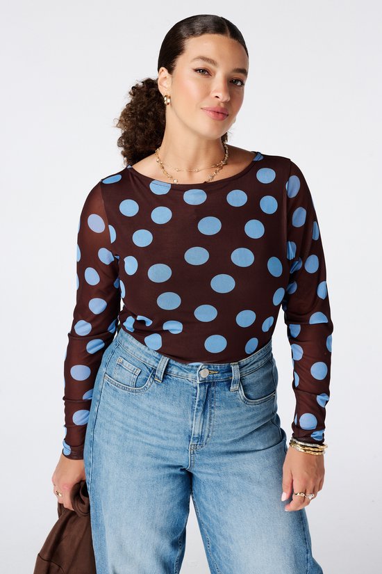 Model draagt een bruine, doorschijnende top met blauwe polkadots en een ronde hals, gecombineerd met een lichtblauwe denim jeans.