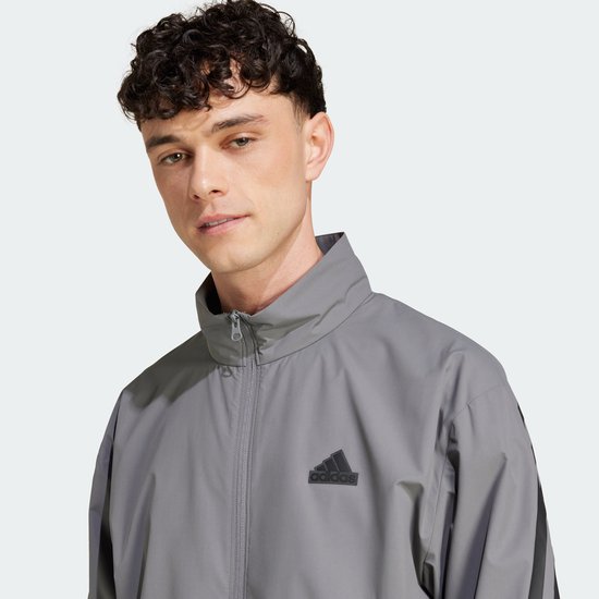 Man met een grijze Adidas trainingsjack met zwarte strepen op de mouwen en een Adidas logo op de borst.