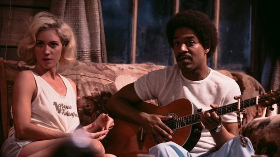 Een vrouw met blond haar draagt een wit tanktop met 'Bayou Boogie' erop, naast een man met afro en gitaar.