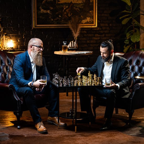 Twee mannen met baard spelen schaak in een luxe kamer. Één man houdt een glas whiskey vast.