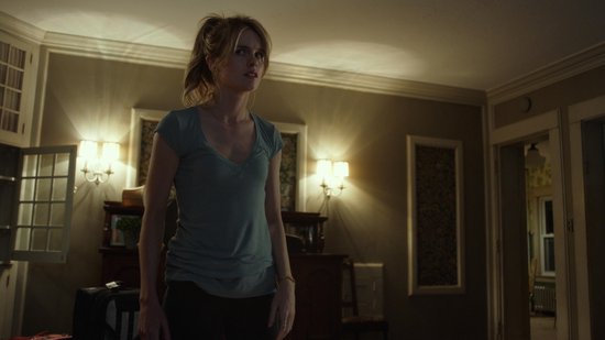 Een vrouw met blond haar in een paardenstaart, in een blauw T-shirt, staat in een kamer met warme verlichting.