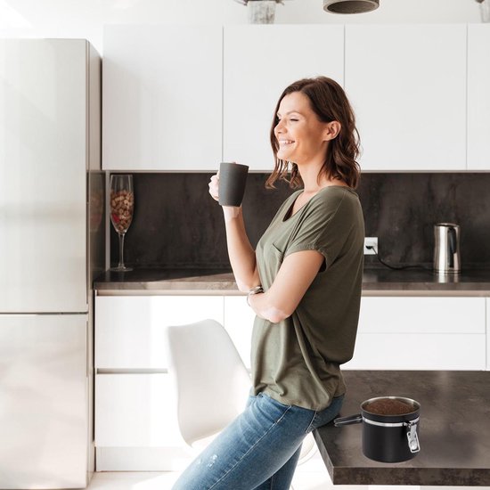 Vrouw in keuken met groene T-shirt en spijkerbroek, drinkt koffie. Op het aanrecht staat een zwarte koffiebus.