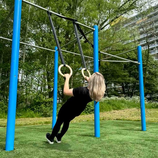 Vrouw in zwarte kleding traint aan houten ringen met zwarte banden aan een rek buiten.