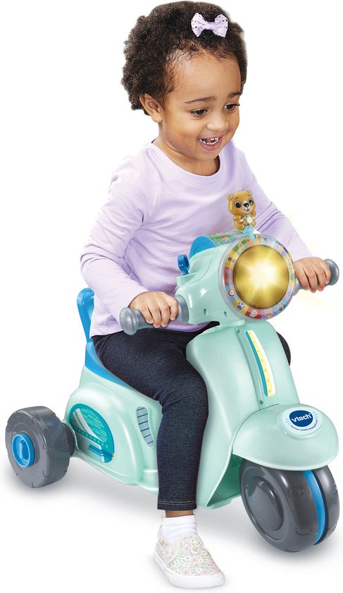 Meisje met krullend haar, lila trui en spijkerbroek zit op een lichtgroene driewielige scooter met blauw zadel en details.