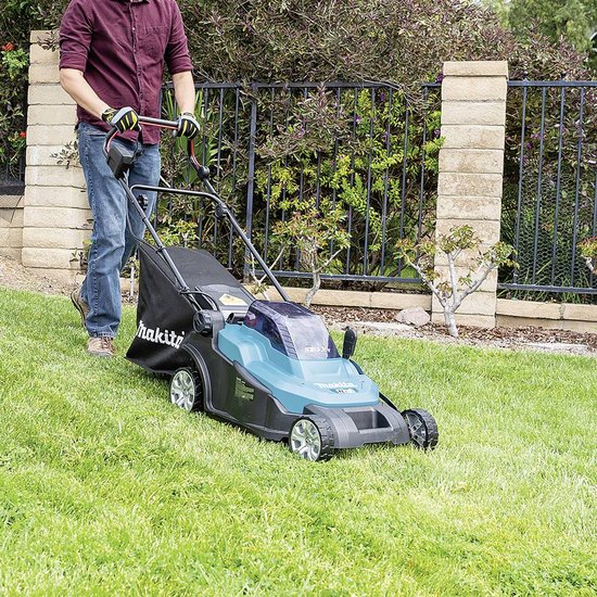Man met grijze trui en gele handschoenen duwt turquoise grasmaaier met opvangzak over gras.