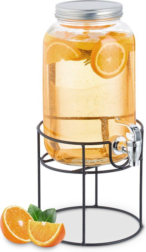 Glazen drankdispenser gevuld met drank en sinaasappelschijfjes, op zwarte standaard. Sinaasappels ernaast.
