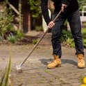 Persoon met gele laarzen gebruikt een houten bezem met metalen borstels op een betegeld terras.