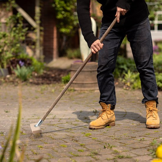 Persoon met gele laarzen gebruikt een houten bezem met metalen borstels op een betegeld terras.