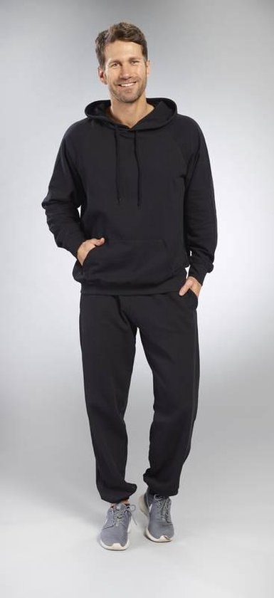 Man draagt een zwarte hoodie met capuchon, buidelzak en zwarte joggingbroek. Handen in de zakken.