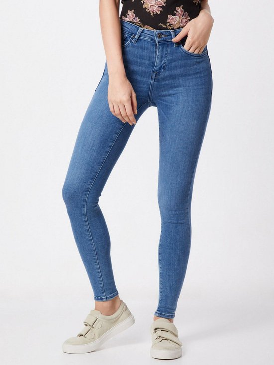 Model draagt een blauwe skinny jeans met middelhoge taille, met witte sneakers.