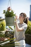 Vrouw met een grijze t-shirt en witte broek, verzorgt witte bloemen in een groene hangpot.