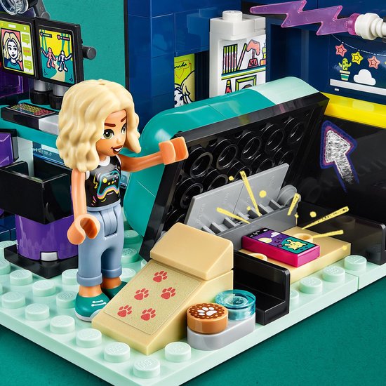 Meisje met blonde krullen in zwart T-shirt met gameprint, grijze broek en sneakers, wijzend naar een spelcomputer.