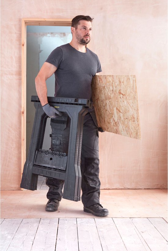 Man met een grijze T-shirt, werkbroek en grijze handschoenen draagt een zwarte, opvouwbare zaagbok.