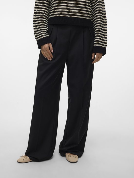 Vrouw draagt een zwarte wijde broek met plooien aan de voorkant, een gestreepte trui en beige platte schoenen.