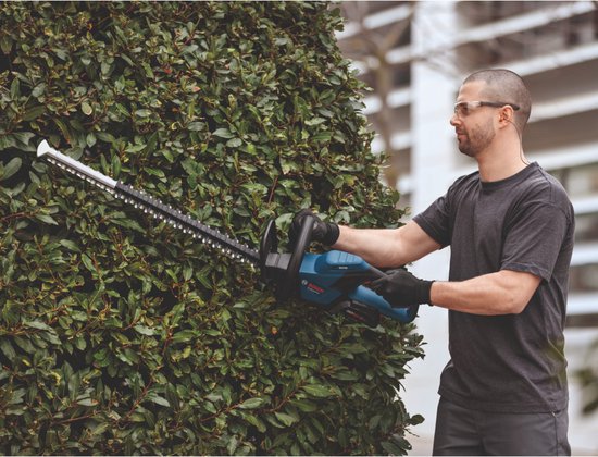 Man met veiligheidsbril en handschoenen snoeit struiken met een blauwe Bosch Professional accu heggenschaar.