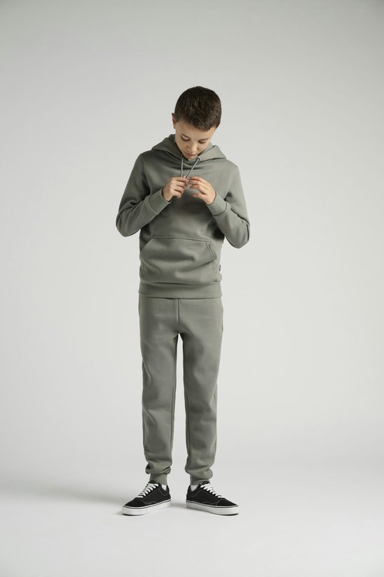 Jongen draagt een olijfgroene joggingbroek met een capuchon. Hij strikt het trekkoord van de capuchon.