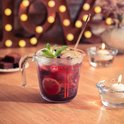 Glazen Pyrex maatbeker gevuld met een rode drank, garnering van munt, limoen en bessen. Twee kaarsen en blokjes chocolade ernaast.