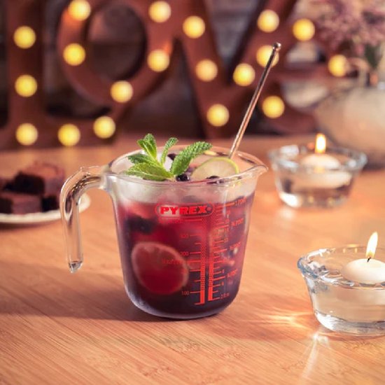 Glazen Pyrex maatbeker gevuld met een rode drank, garnering van munt, limoen en bessen. Twee kaarsen en blokjes chocolade ernaast.