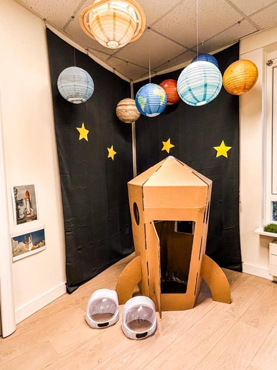 Kamer met een kartonnen raket, twee astronautenhelmen, planeetdecoratie aan het plafond en een zwarte achtergrond.