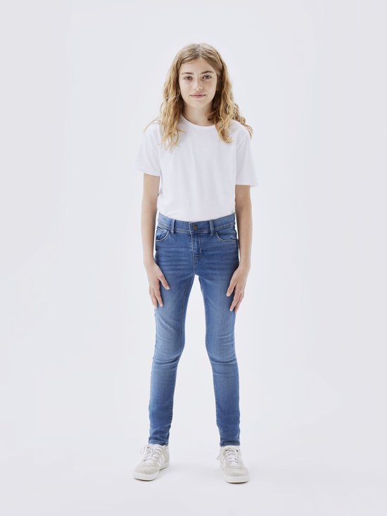 Meisje met lang blond haar, staand in een witte T-shirt en een spijkerbroek met skinny fit.