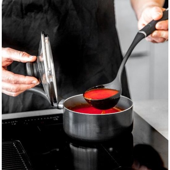 Une main sort une louche de sauce rouge d'une casserole noire sur une plaque de cuisson.