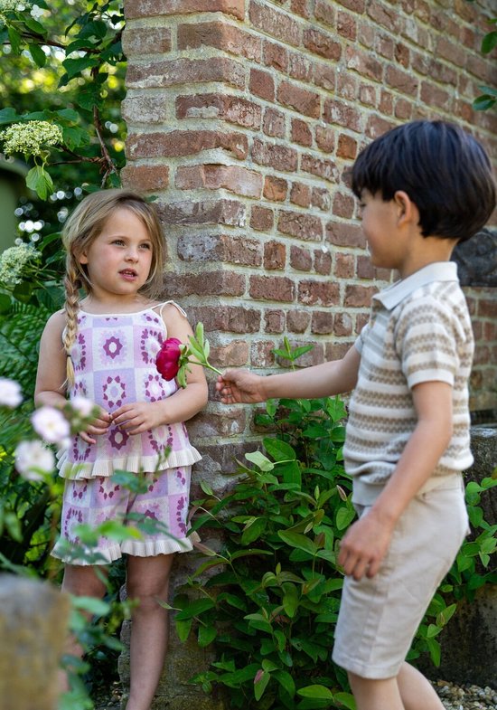 Meisje met blonde vlechtjes, lila top met bloemenprint en ruches, bij een stenen muur.