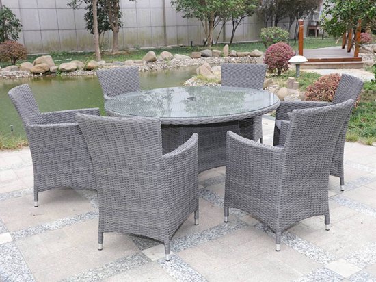 Ronde grijze wicker eettafel met glazen blad, omringd door zes grijze wicker stoelen met armleuningen.