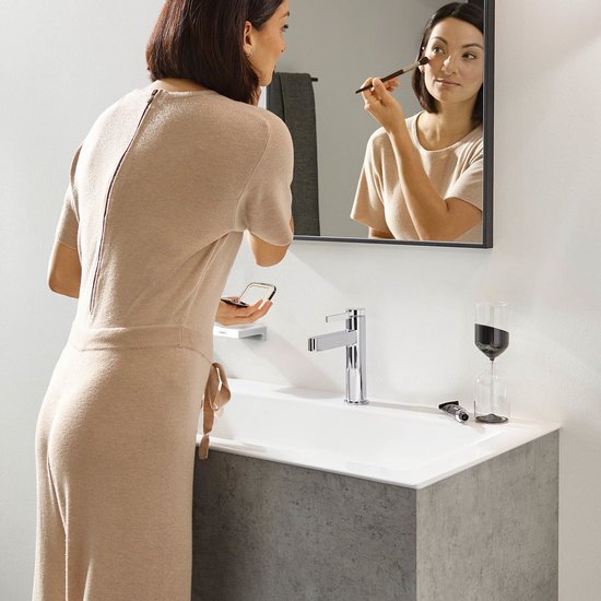 Een vrouw met een beige jurk brengt make-up aan bij een spiegel boven een chromen wastafelkraan.