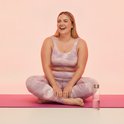 Vrouw in roze tie-dye sportbeha en legging, zittend op een yogamat met een fles water.