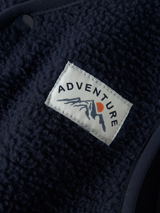 Close-up van het 'ADVENTURE' label op een donkerblauw teddy gilet met bergillustratie.
