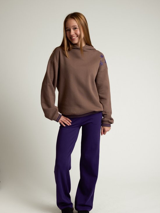 Model met lachend gezicht draagt een bruine hoodie met sterren en een paarse broek.