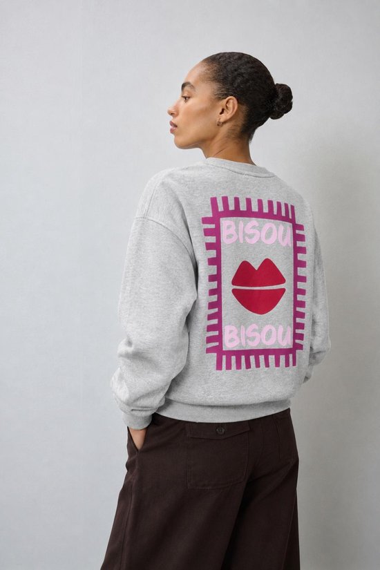 Model draagt lichtgrijze sweater met rode lip en 'BISOU BISOU' opdruk op de rug.