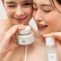 Twee vrouwen met een glimlach houden een potje en flesje Beauty of Joseon vast. De een knipoogt.