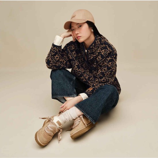 Model draagt een bruin vest met patroon, spijkerbroek, beige pet en UGG Lowmel sneakers in lichtbruin.