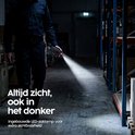 Beveiliger loopt door donker magazijn met ingeschakelde LED-zaklamp op portofoon.