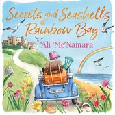 Boekomslag 'Secrets and Seashells at Rainbow Bay' door Ali McNamara. Een blauwe auto vol koffers rijdt naar een kasteel, met bloemen, schelpen en een regenboog.