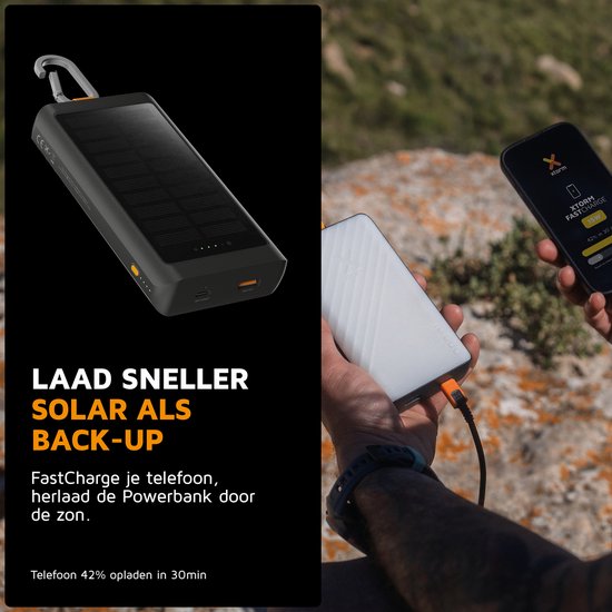 Zwarte Xtorm powerbank met zonnepaneel, karabijnhaak en indicatielampjes, naast een hand die een smartphone oplaadt.