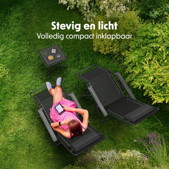Een vrouw in een roze jurk ontspant op een zwarte ligstoel in de tuin, met een tablet. Naast haar staat een bijzettafel.