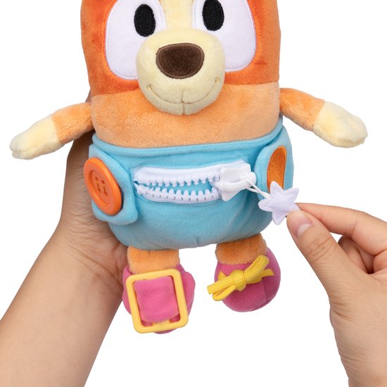 Jouet en peluche orange avec un pantalon bleu, une fermeture éclair, des boutons et des chaussures. Une main ouvre la fermeture éclair.