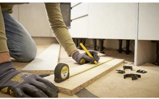 Man met grijze handschoenen legt een Stanley rolbandmaat naast een stuk hout en enkele klemmen.