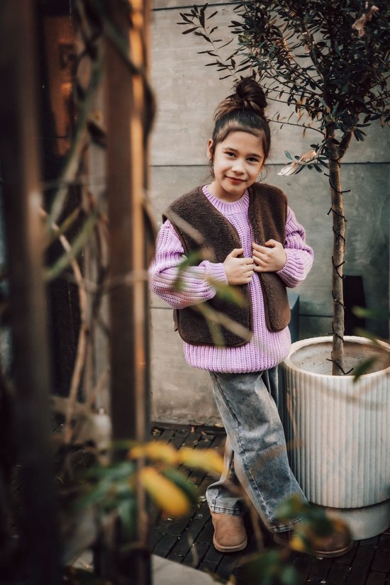Meisje met opgestoken haar draagt een bruin teddy fleece gilet over een paarse gebreide trui.