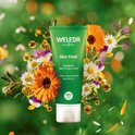 WELEDA Skin Food tube te midden van bloemen, bladeren en oliebolletjes. Groene tube met witte dop. "Voedende alles-in-één crème. Crème nourrissante tout-en-un."