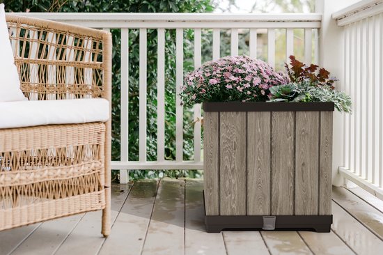 Vierkante grijze plantenbak met houtlook, met bloemen, naast een rotan stoel op een balkon.